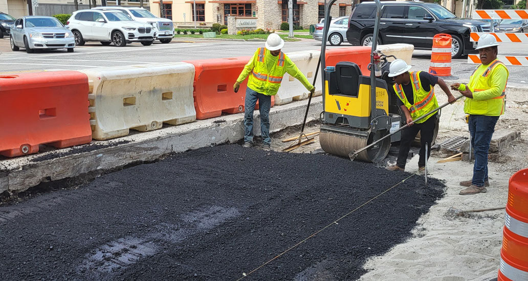 Construction workers spreading and use a steel roller to flatten the freshly placed black asphalt surface on the corner of Westheimer and Weslayne.